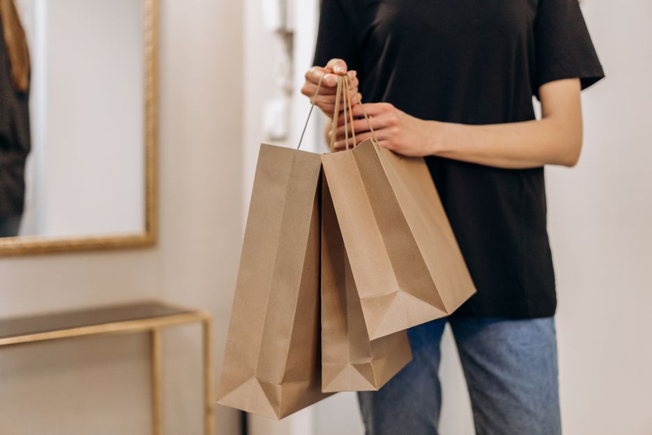 A person standing indoors holding three brown paper shopping bags in one hand, wearing casual clothing.